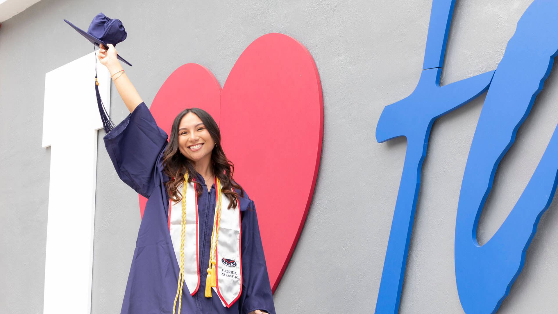 Graduate in cap and gown celebrating in front of a colorful 'I ❤️ FAU' wall, representing Florida Atlantic University.