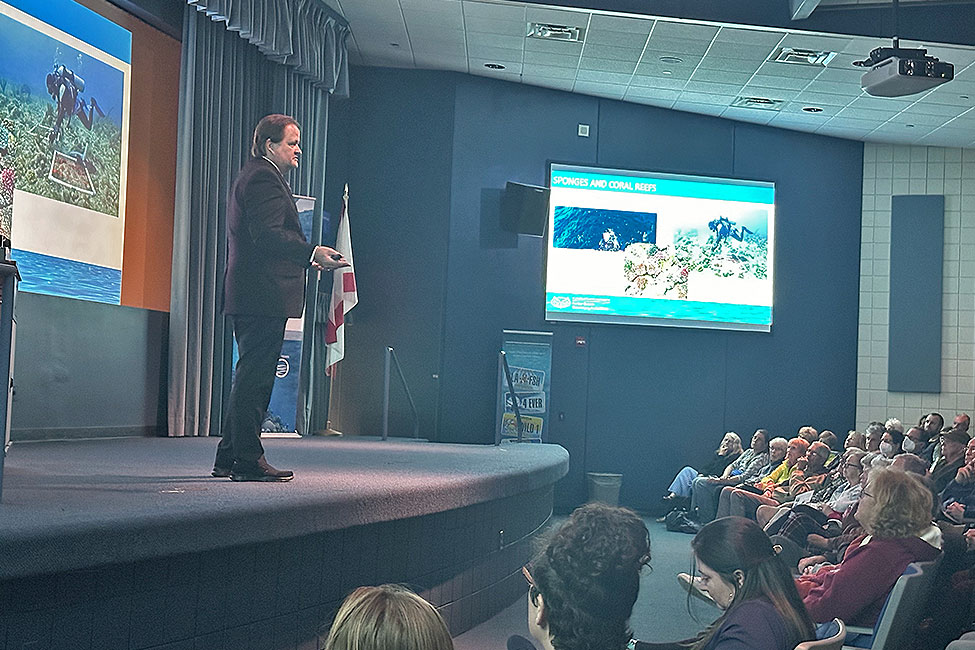 Man stands on a stage presenting in front of an audience in a darkened auditorium. Large screens display underwater scenes.