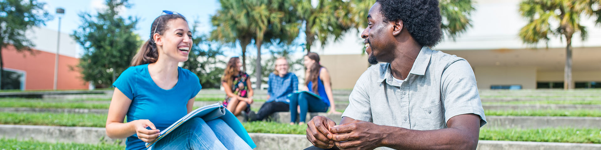 Students sitting on the steps in front of the College of Arts and Letters. Two students are in the foreground talking and laughing. Three more students are in the background.