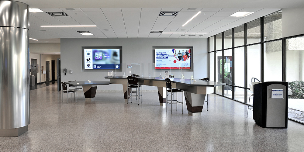 Student Union lobby with a spacious, modern design, featuring high-top tables with stools, digital campus display screens, and floor-to-ceiling windows providing natural light and outdoor views
