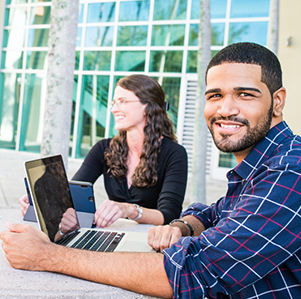 Male and female student sitting at an outdoor table on FAU campus on their laptops. The male is looking at the camera and smiling.