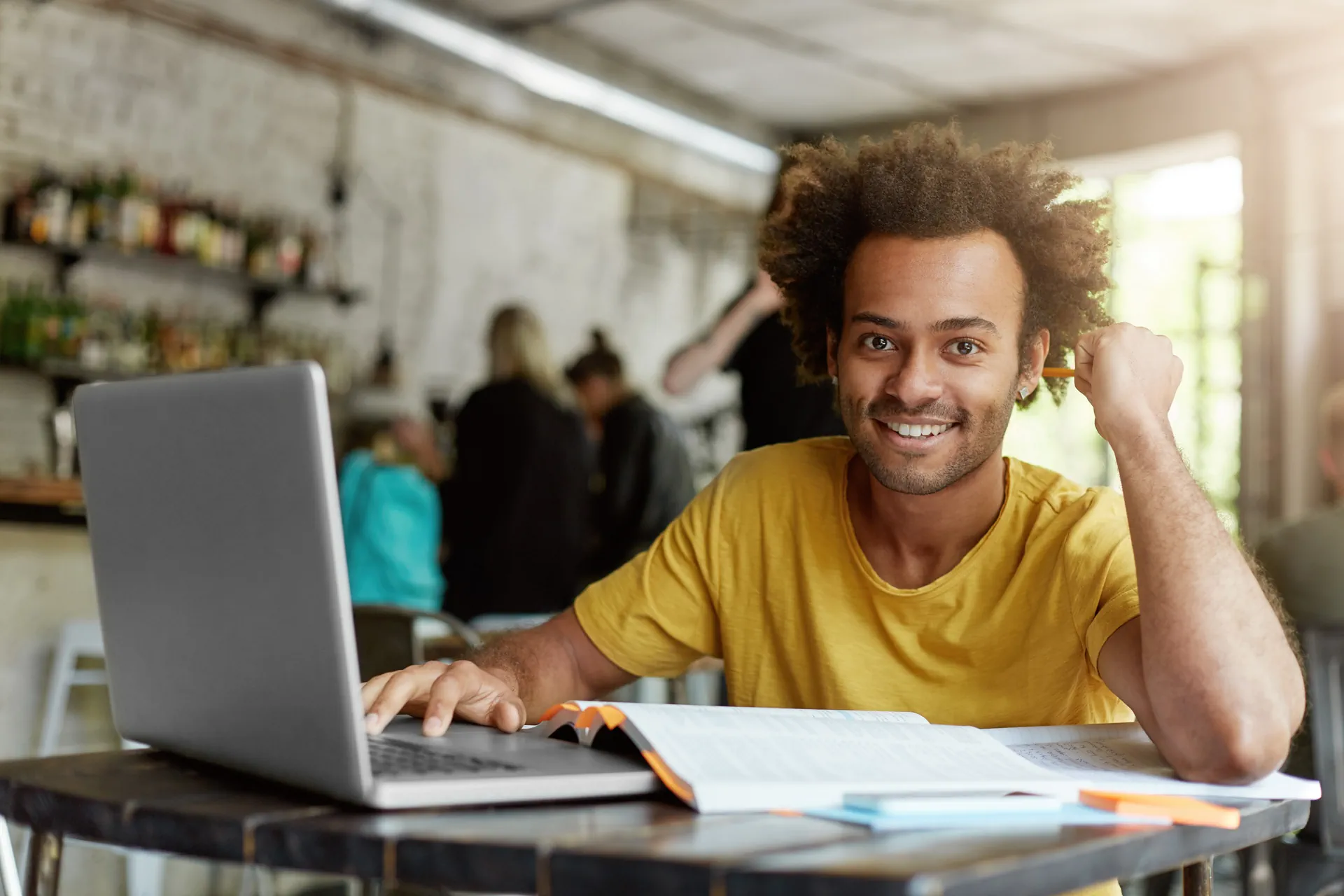 A smiling man in a yellow shirt works on a laptop with an open book